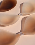 Product shot of reusable and sticky Dark tint, Light tint, and Medium tint Silicone Stick-on Bras laying flat against light pink background, close-up shot showing the clasps and thin edges on each bra