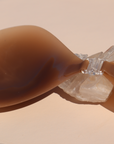 Product shot of sticky and reusable Dark tint Silicone Stick-on Bra laying against a silica crystal and light pink background, close-up front shot of clasp and razor thin edge
