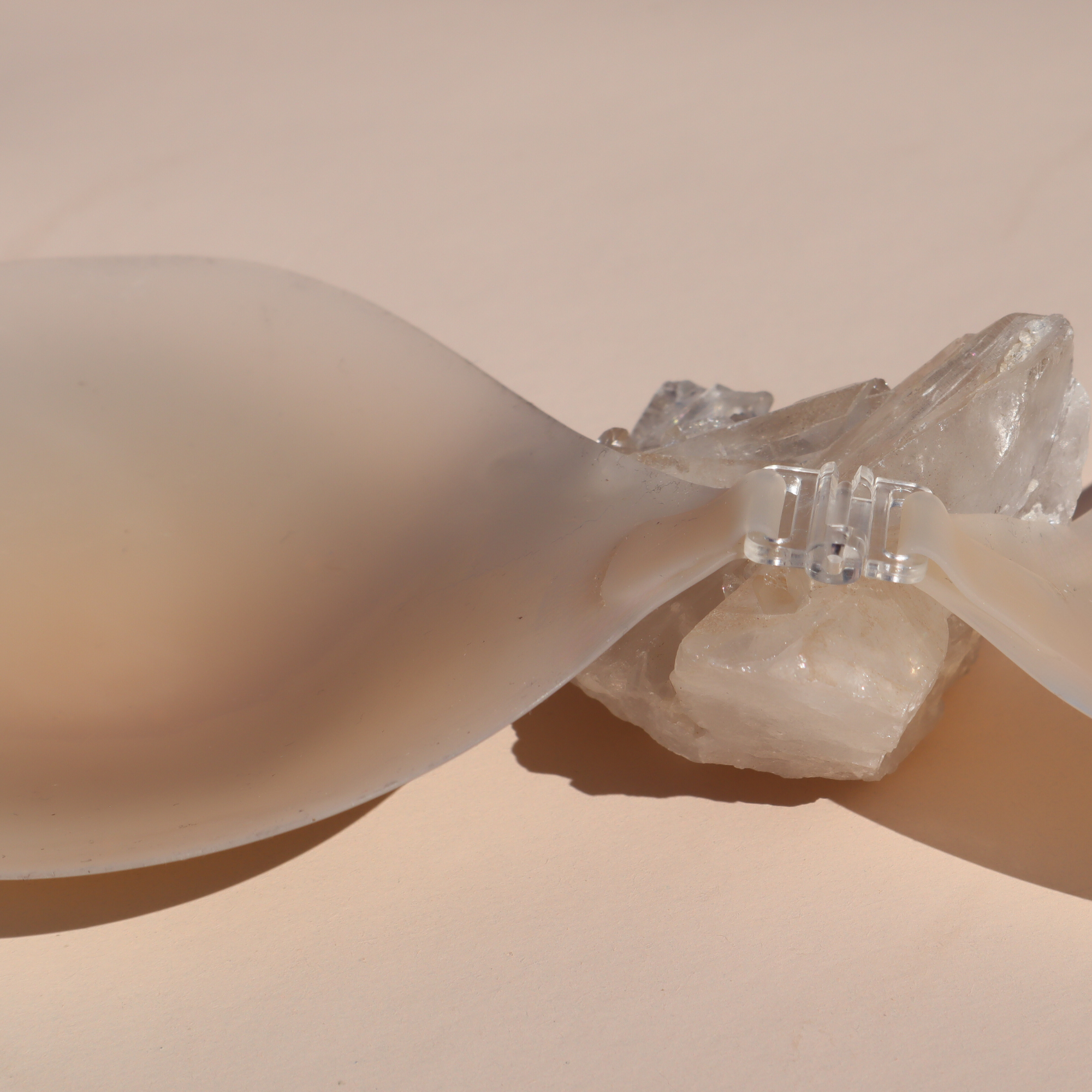 Product shot of reusable and sticky Light tint Silicone Stick-on Bra laying against silica crystal and light pink background, close-up shot showing the clasp and razor thin edges of the Silicone Stick-on Bra