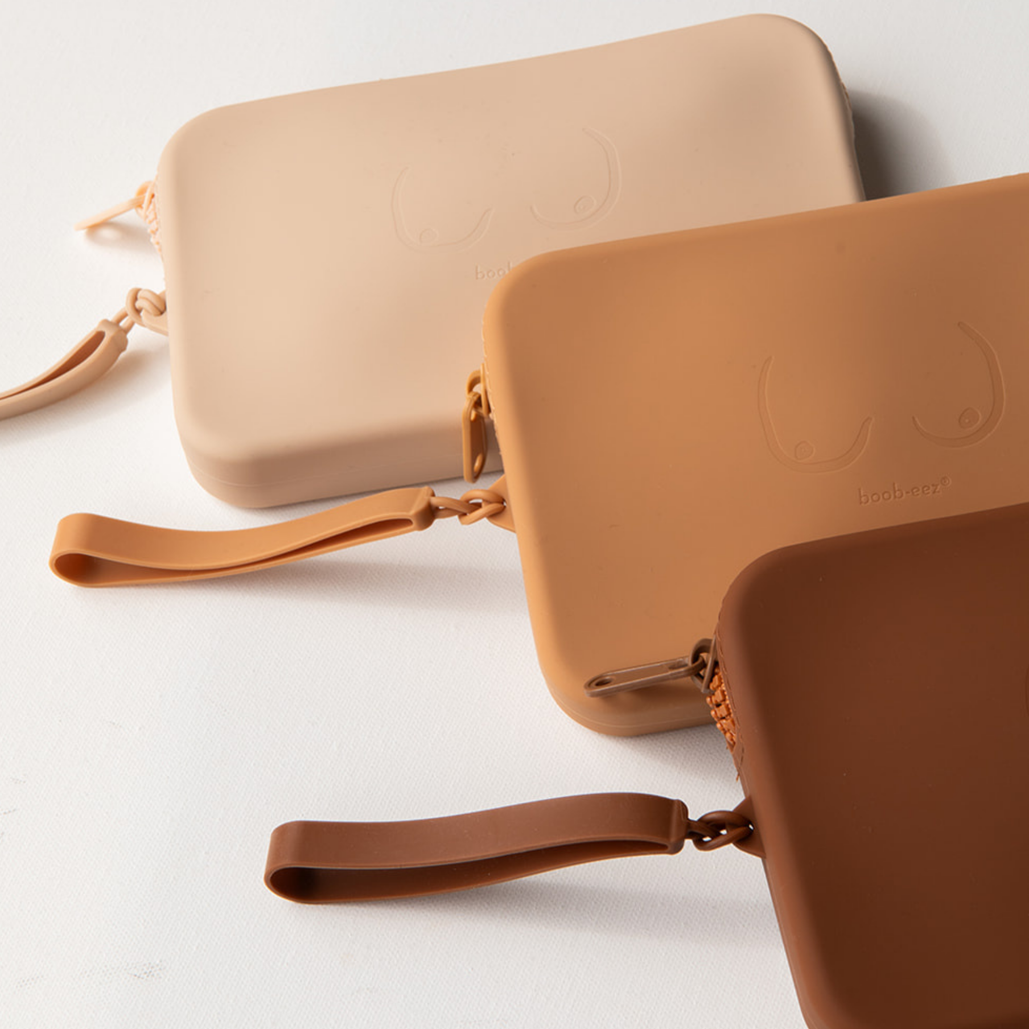 Product shot of durable Silicone Travel Cases for Silicone Stick-on Bra in Light Nude, Medium Nude, and Dark Nude, against a white background showing the Boob-eez branded detail and removable strap