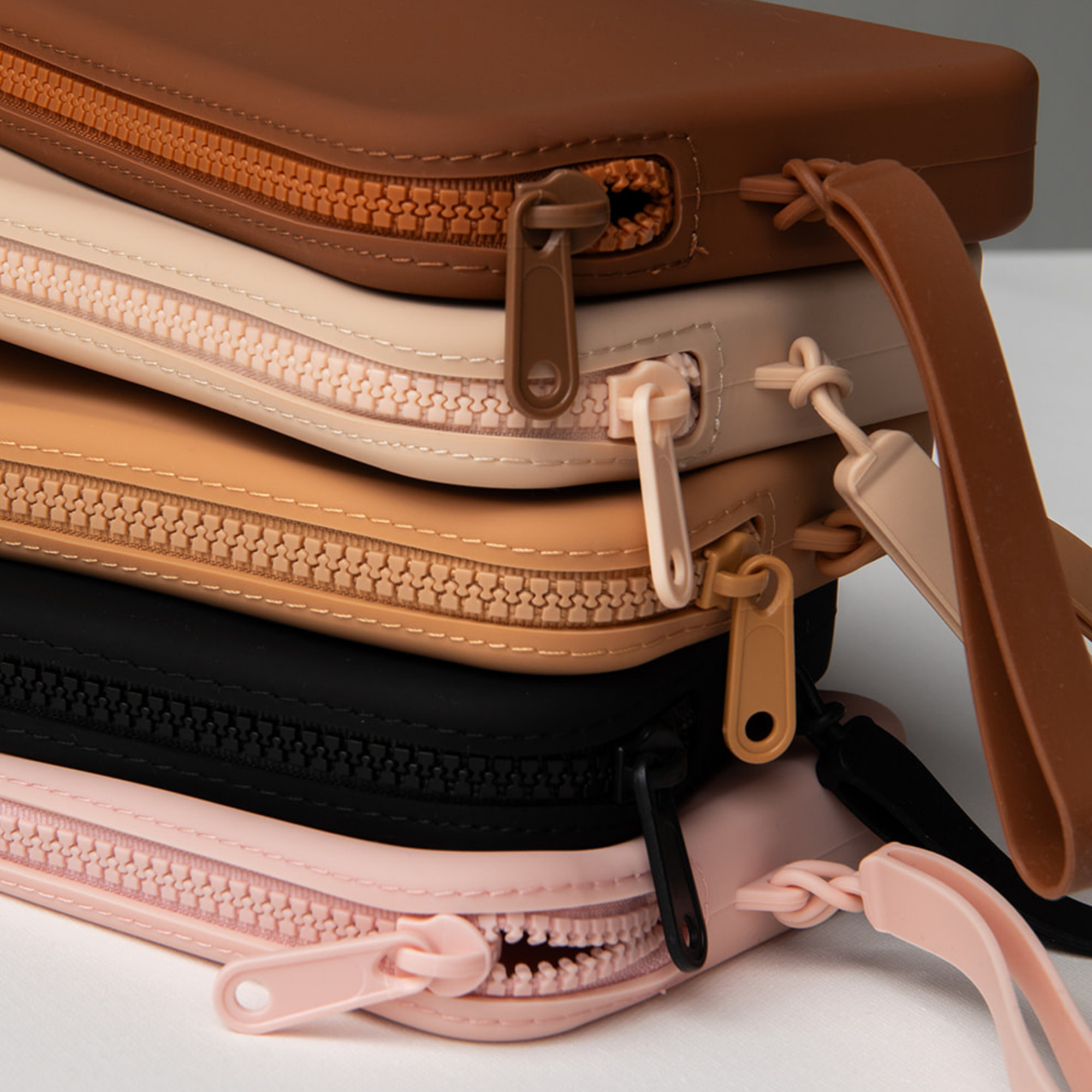 Product shot of a stack of reusable Silicone Travel Cases for Silicone Stick-on Bra in Dark Nude, Light Nude, Medium Nude, Black, and Dusty Pink, showing the zipper detail and removable strap
