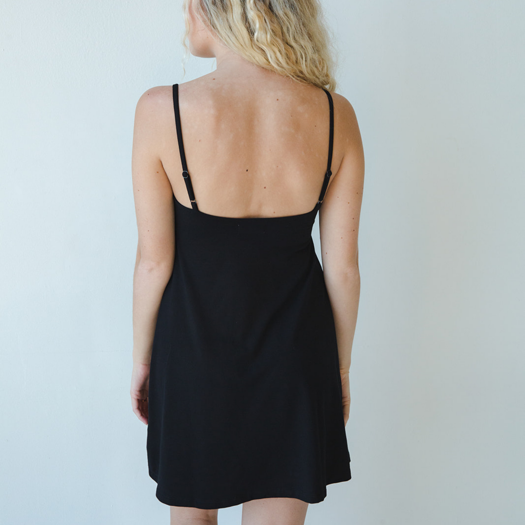 Product shot of NŪD Apparel Betty Swinging Slip Dress, thin adjustable strap, loose style and with shelf bra, made of bamboo fabric produced in California, in Black on fair-skinned model, back shot
