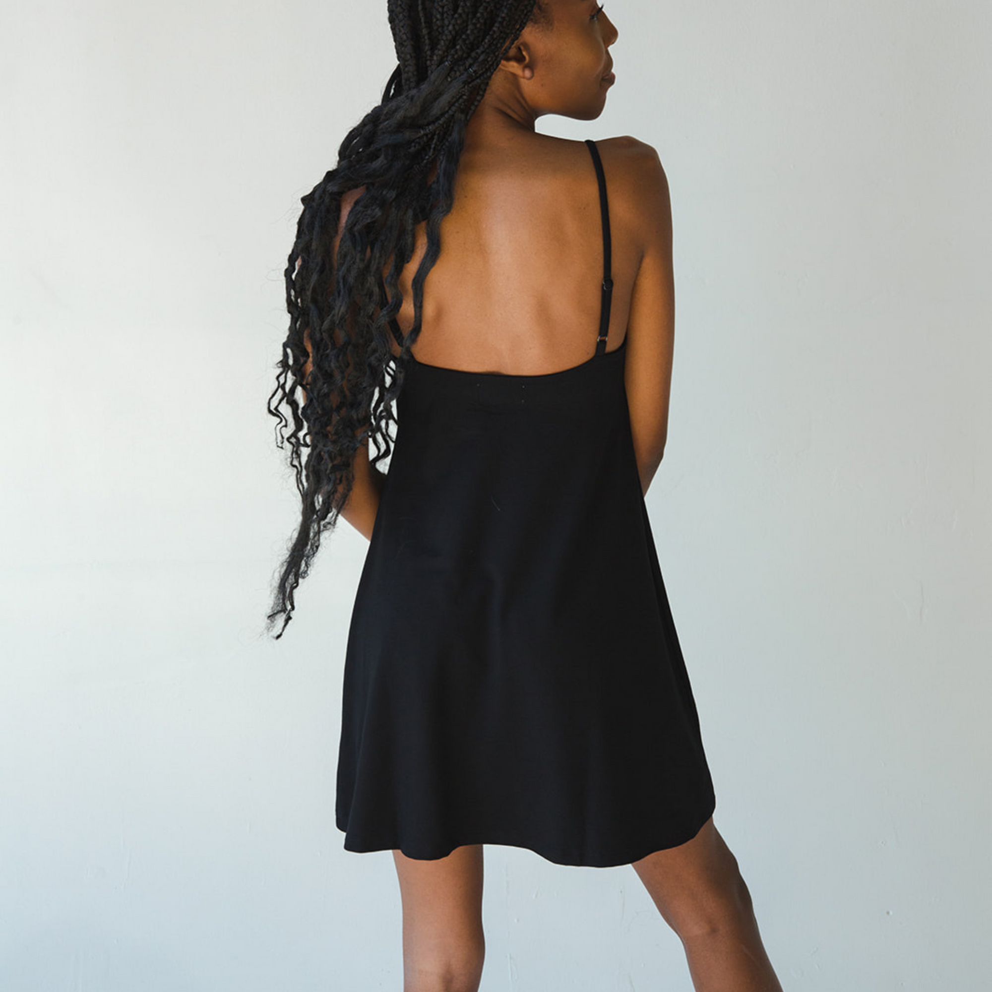 Product shot of NŪD Apparel Betty Swinging Slip Dress, thin adjustable strap, loose style and with shelf bra, made of bamboo fabric produced in California, in Black on dark-skinned model, back shot