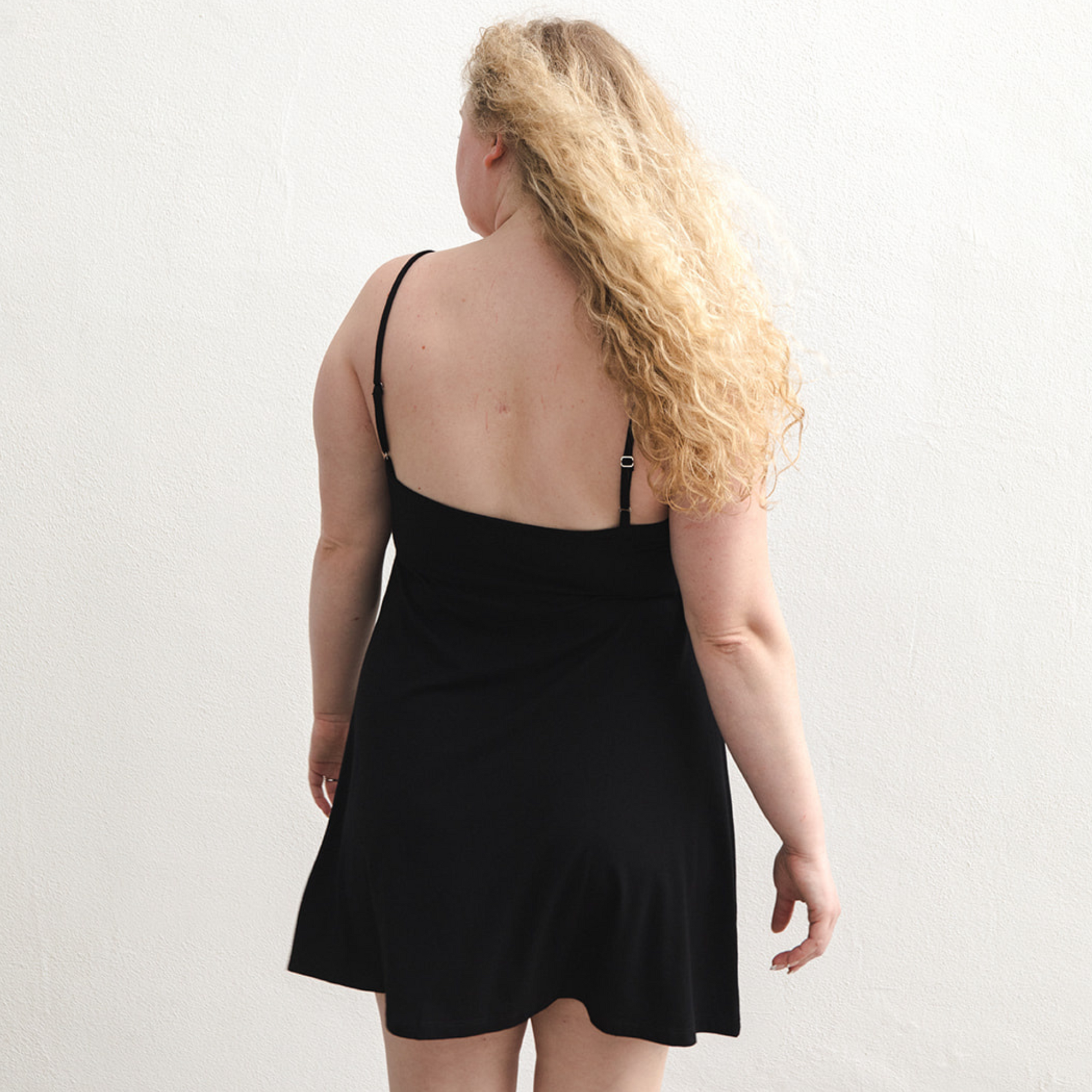 Product shot of NŪD Apparel Betty Swinging Slip Dress, thin adjustable strap, loose style and with shelf bra, made of bamboo fabric produced in California, in Black on fair-skinned model, back shot