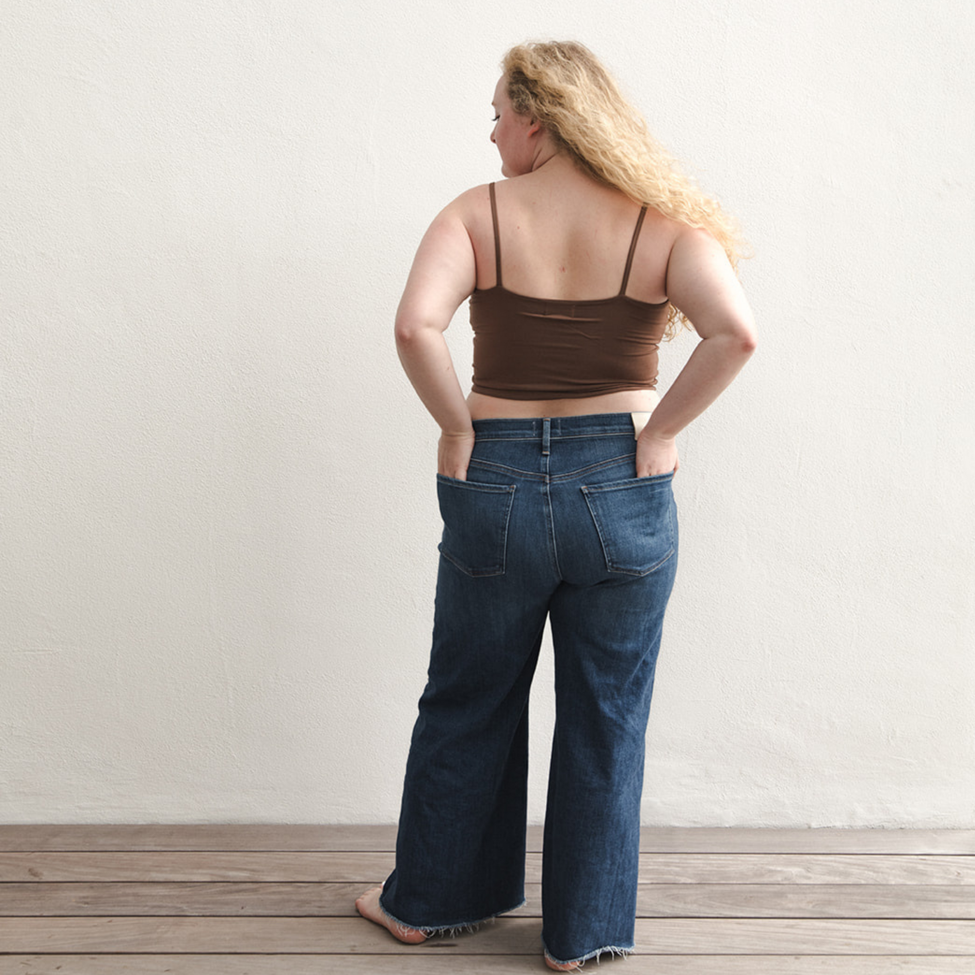 Product shot of NŪD Apparel Kyla Double Layered Crop top, thin strap and seamless, made of bamboo fabric produced in California, in Dark Nude on fair-skinned model paired with dark-wash jeans, back wide shot