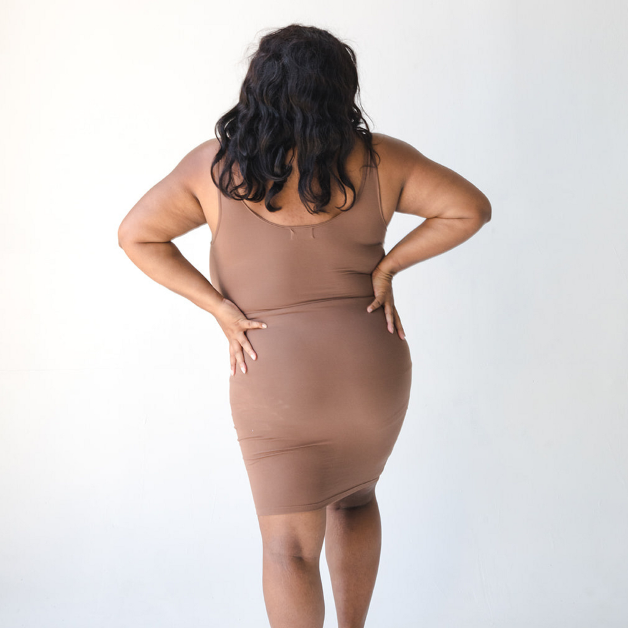 Product shot of NŪD Apparel Tayler Slip Dress, wide strap, fitted style and seamless, made of bamboo fabric produced in California, in Dark Nude on dark-skinned model, back shot