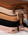 Product shot of a stack of reusable Silicone Travel Cases for Silicone Stick-on Bra in Dark Nude, Light Nude, Medium Nude, Black, and Dusty Pink, showing the zipper detail and removable strap