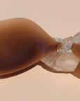 Product shot of sticky and reusable Dark tint Silicone Stick-on Bra laying against a silica crystal and light pink background, close-up front shot of clasp and razor thin edge