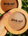 Product shot of a hand holding a Boob-eez Bamboo Compact with Dark tint Headlight Hiders inside and a Bamboo Compact with Medium tint nipple covers inside with natural green in the background, close-up shot showing the beautiful wood grain of the bamboo and the transparent window to show the sticky nipple covers inside