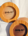 Product shot of a hand holding a Boob-eez Bamboo Compact with Dark tint Headlight Hiders inside and a Bamboo Compact witjh Light tint nipple covers inside with sticky nipple covers in the background, close-up shot showing the beautiful wood grain of the bamboo