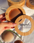 Product shot of a hand holding a Boob-eez Bamboo Compact with Dark tint Headlight Hiders inside with Bamboo Compacts and nipple covers in the background, close-up shot showing the beautiful wood grain of the bamboo and the magnetic closure