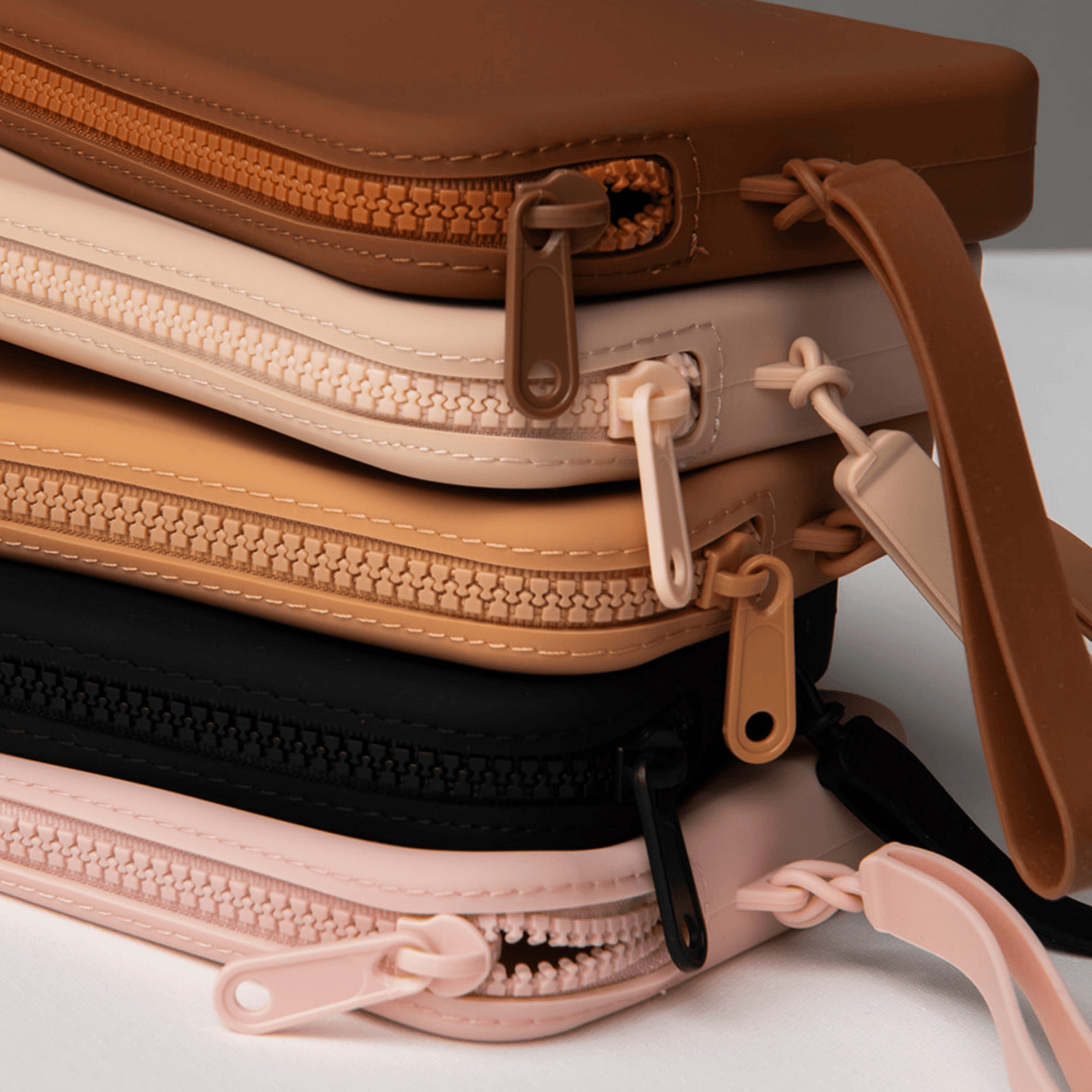 Product shot of a stack of reusable Silicone Travel Cases for Silicone Stick-on Bra in Dark Nude, Light Nude, Medium Nude, Black, and Dusty Pink, showing the zipper detail and removable strap