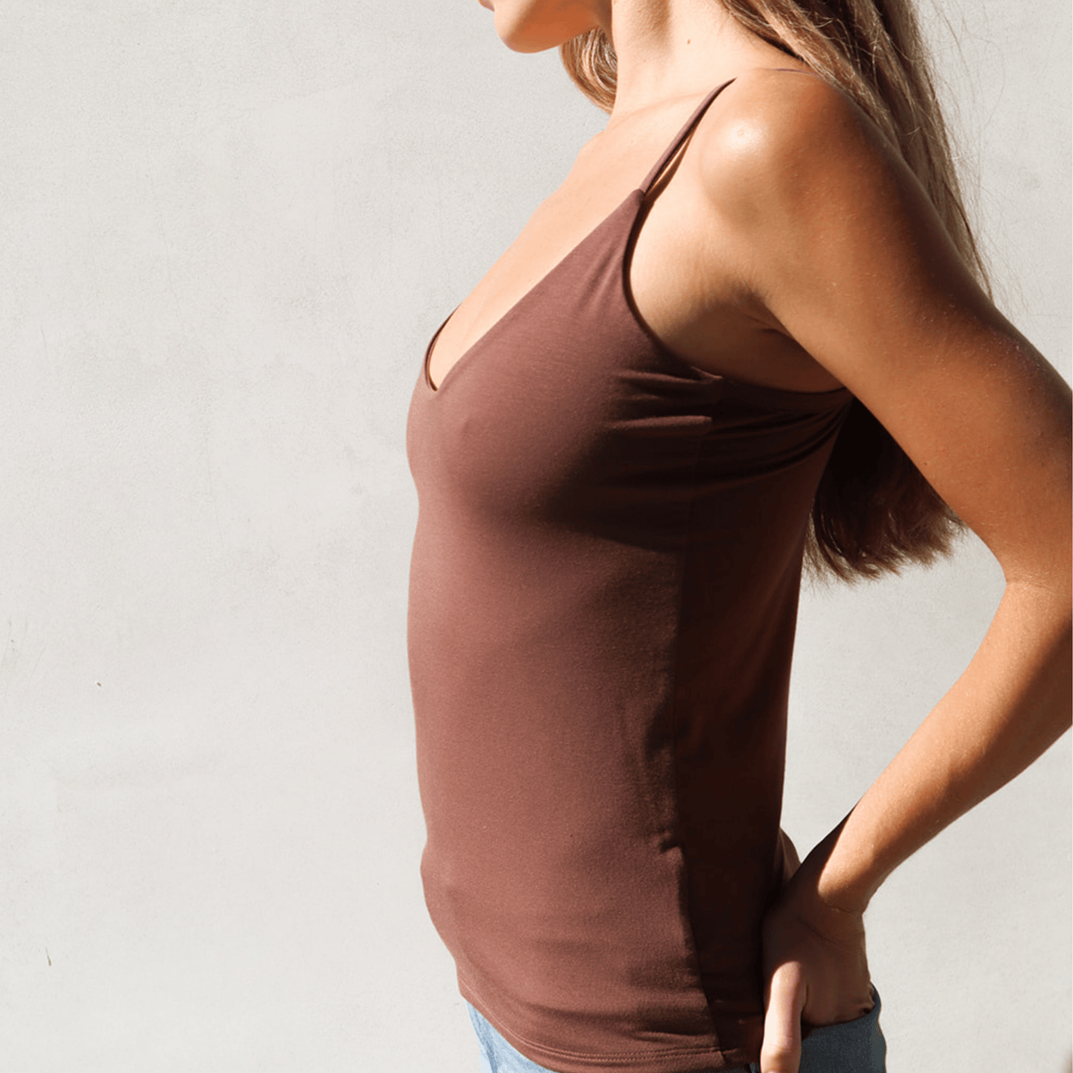 Product shot of NŪD Apparel Nesha Double Layered Tank, thin strap, long style and seamless, made of bamboo fabric produced in California, in Dark Nude on fair-skinned model paired with light-wash jeans, side shot